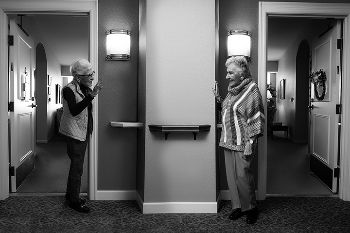 Two older women stand in a hallway with wall sconce lighting as they greet each other from their respective neighboring apartment doors. Black and white photo.