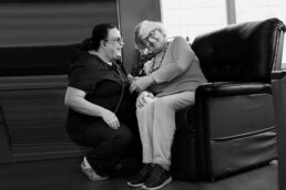 A female caregiving wearing scrubs and a holding a stethoscope, kneels in front of an older woman in a lounge chair as they grasp hands and smile. Black and white photo.