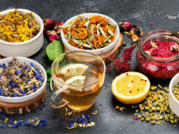 An array of colorful dried herbs in bowls on a dark surface, accompanied by a glass of herbal tea with lemon, ginger, and a halved lemon, conveying a fresh, aromatic mood.