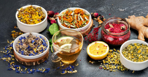An array of colorful dried herbs in bowls on a dark surface, accompanied by a glass of herbal tea with lemon, ginger, and a halved lemon, conveying a fresh, aromatic mood.