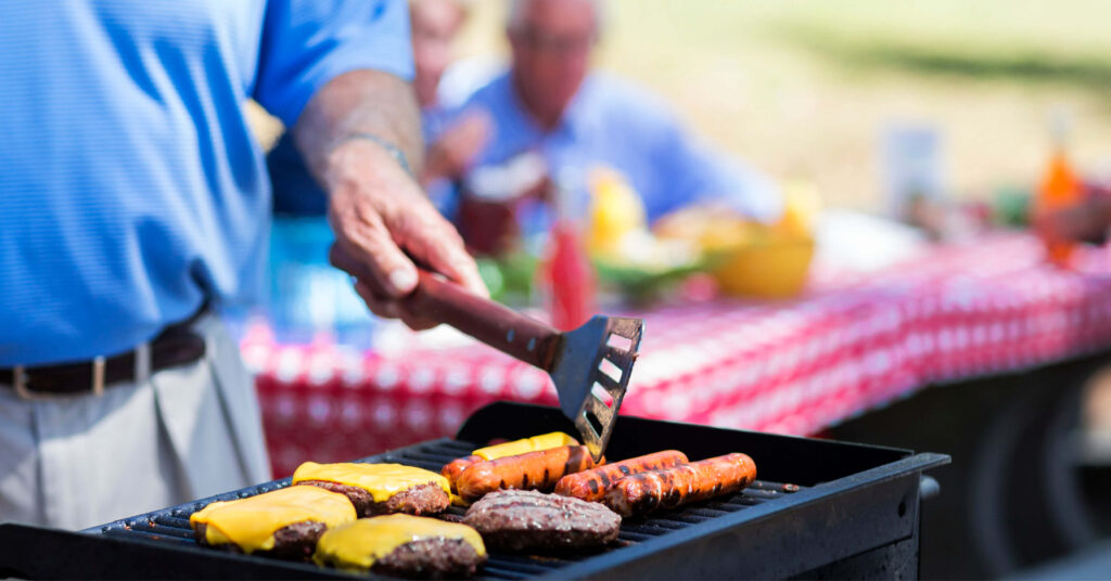 A person grills burgers with melted cheese and sausages on a barbecue. In the background, people sit at a picnic table with a red checkered cloth.