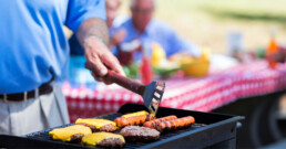 A person grills burgers with melted cheese and sausages on a barbecue. In the background, people sit at a picnic table with a red checkered cloth.