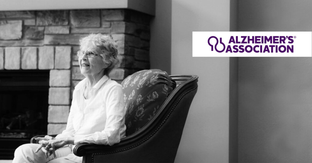Elderly woman smiling while seated in an armchair, set against a stone fireplace backdrop. Alzheimer's Association logo in the top right corner.