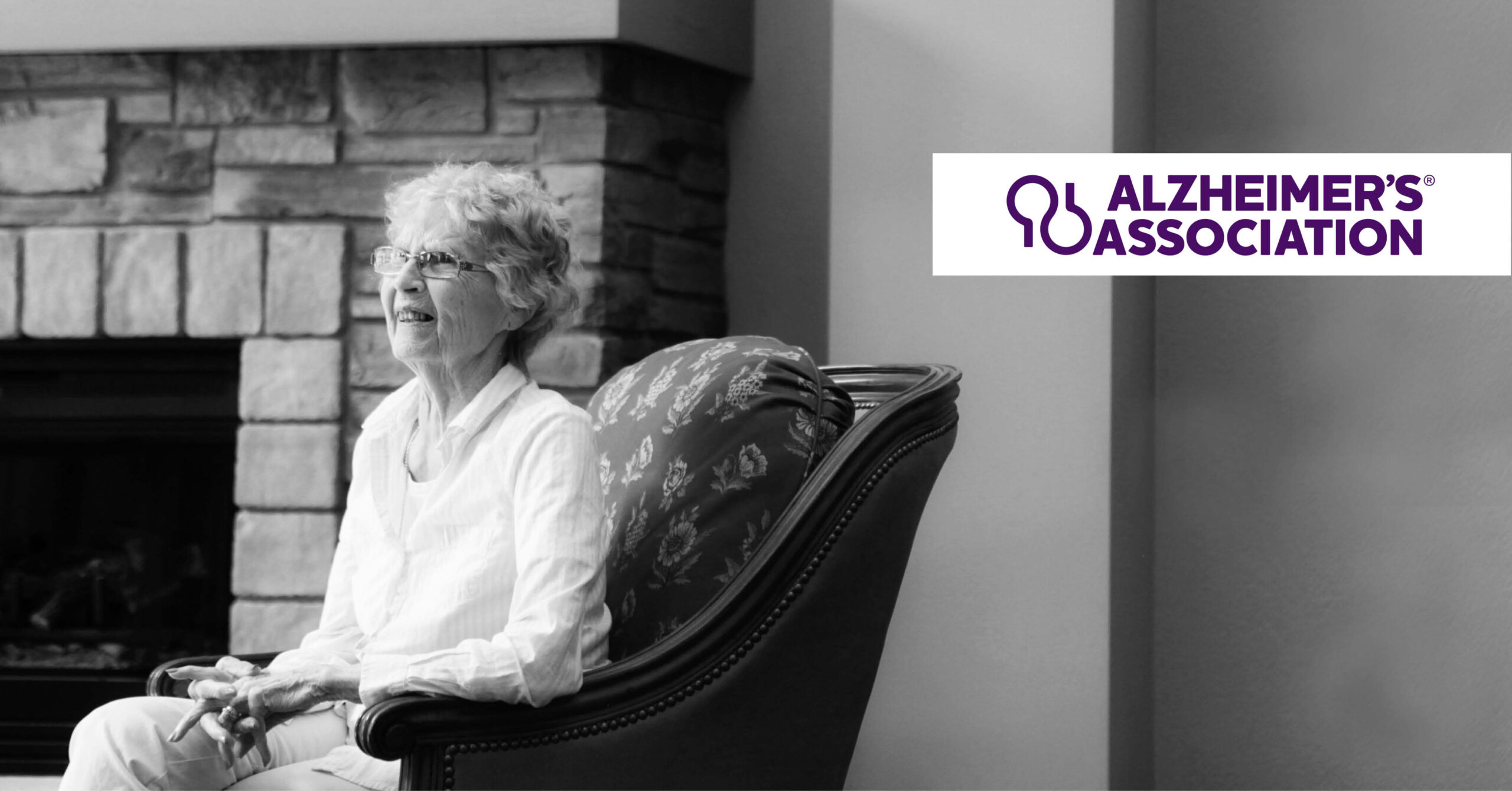 Elderly woman smiling while seated in an armchair, set against a stone fireplace backdrop. Alzheimer's Association logo in the top right corner.
