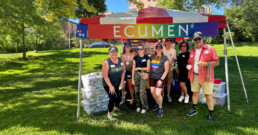A group of eight people smiling under a colorful Pride tent with 
