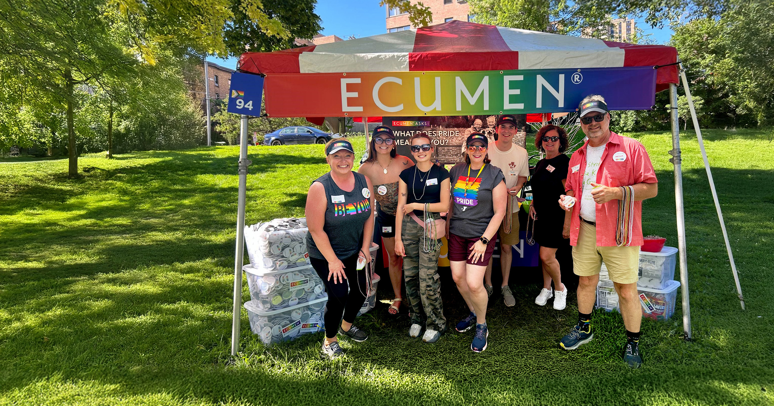 A group of eight people smiling under a colorful Pride tent with "Ecumen" signage in a sunny park setting, conveying a cheerful and inclusive atmosphere.