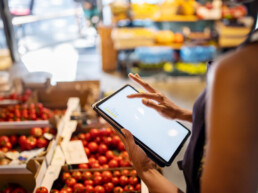 A person using a tablet stands beside boxes of fresh tomatoes in a grocery store. The scene is vibrant and busy, suggesting a modern shopping experience.