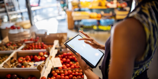 A person using a tablet stands beside boxes of fresh tomatoes in a grocery store. The scene is vibrant and busy, suggesting a modern shopping experience.