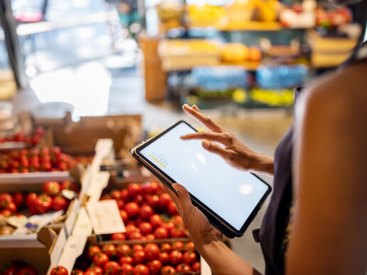 A person using a tablet stands beside boxes of fresh tomatoes in a grocery store. The scene is vibrant and busy, suggesting a modern shopping experience.