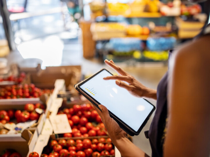 A person using a tablet stands beside boxes of fresh tomatoes in a grocery store. The scene is vibrant and busy, suggesting a modern shopping experience.
