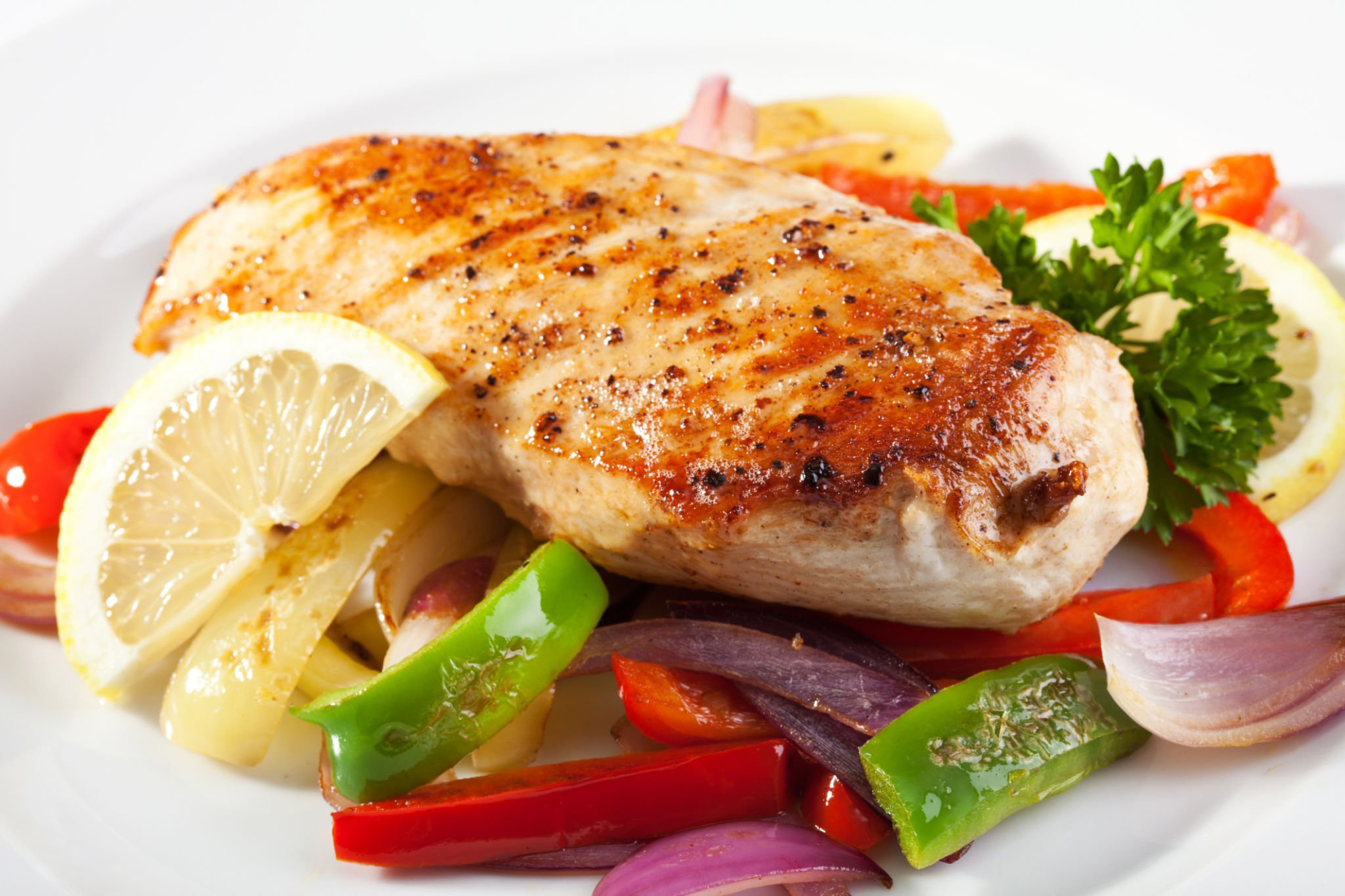 Grilled chicken breast on a white plate, garnished with lemon slices, parsley, and colorful sautéed bell peppers and onions. The dish looks fresh and appetizing.
