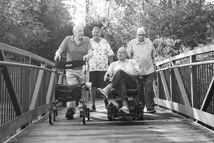 Black and white photo of four older adults crossing a wooden bridge with iron railings. One pushes a wheeled walker, another drives a motorized chair, and two walk behind.