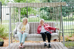 Two older woman outside sitting on an upholstered bench swing as they talk and laugh.
