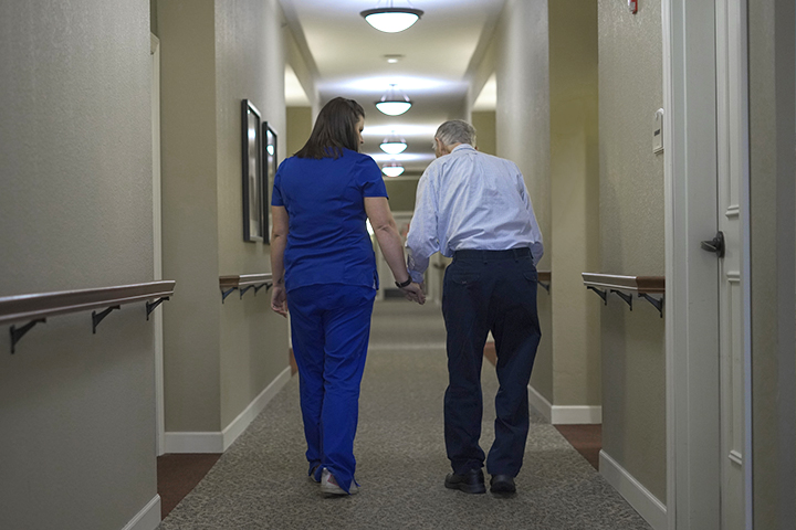 A female nurse walking with an older man down a hallway and providing assistance.