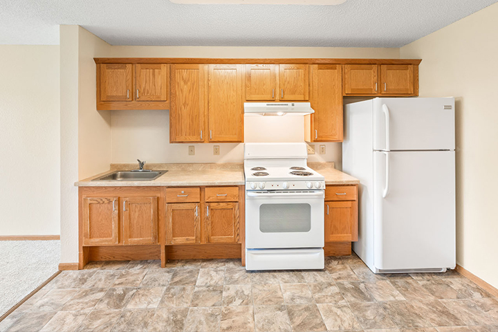 Interior view of assisted living apartment kitchen with wood cabinetry, white appliances and tile flooring that meets beige carpeting.