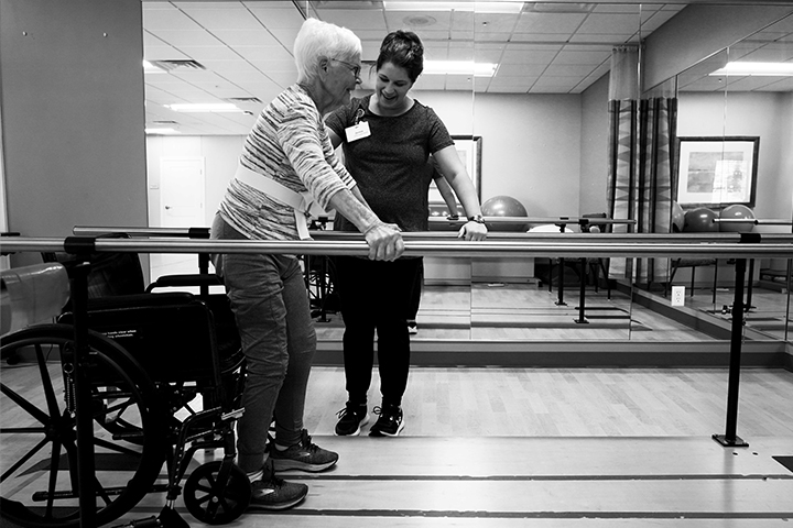 Black and white photo of an older woman using parallel bars for support during physical therapy, assisted by a healthcare professional.