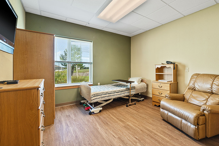 Interior view of a long-term care suite, featuring wood flooring, a large window, wooden cabinets, wall-mounted TV, upholstered lounge chair, and an adjustable hospital-style bed with linens.