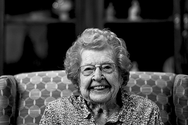 Black and white photo of an older woman wearing glasses and sitting on a lounge chair, smiles brightly at the camera.