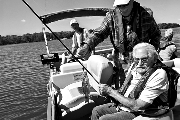 Black and white photo of an older man sitting in a fishing boat on a lake while holding a rod and reel to show off his catch of the day to others on the boat.