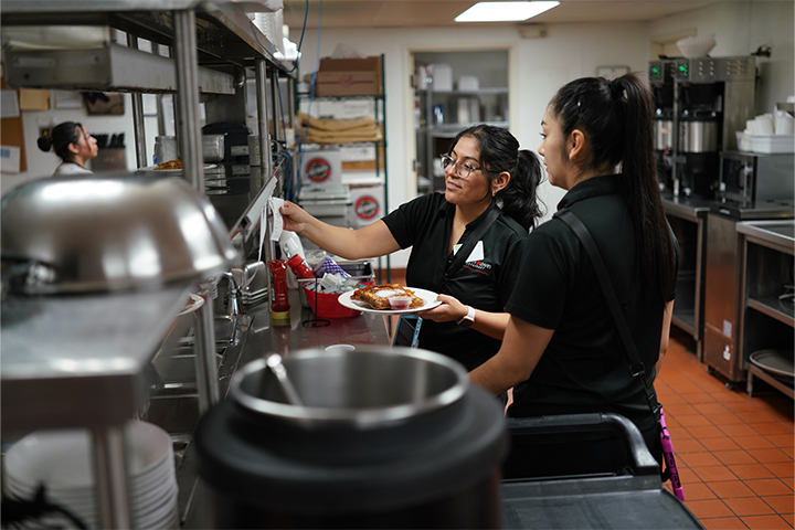 Two young female dining staff in a commercial kitchen with their hair in ponytails as they plate a meal.