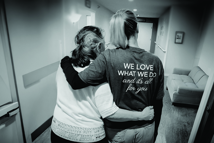 Black and white photo from behind a younger woman and an older woman with an arm around each other as they walk down a hallway. The shirt on the younger woman reads "We love what we do and it's all for you."