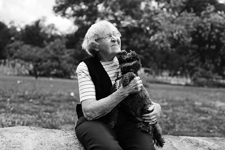 Black and white photo of an older woman kneeling down to embrace a small poodle as it licks her chin.