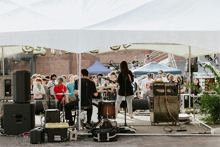Two musicians under a white tent shelter facing a crowd of people.