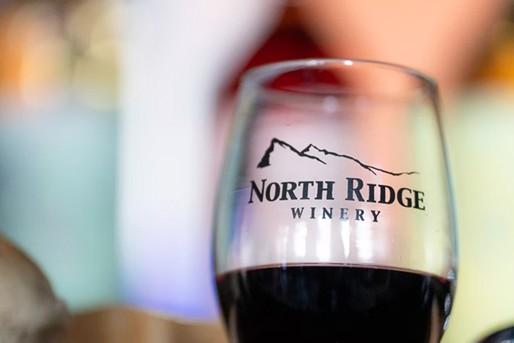 A close-up image of a wine glass holding red wine. The glass has a logo on it that reads North Ridge Winery.