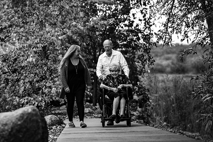 Black and white photo of a young woman walking on a paved path next to an older gentleman who is pushing an older woman in a wheel chair.