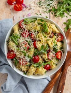 A vibrant bowl of tortellini salad with cherry tomatoes, arugula, and shredded cheese, set on a wooden table with a blue napkin and wooden utensils.