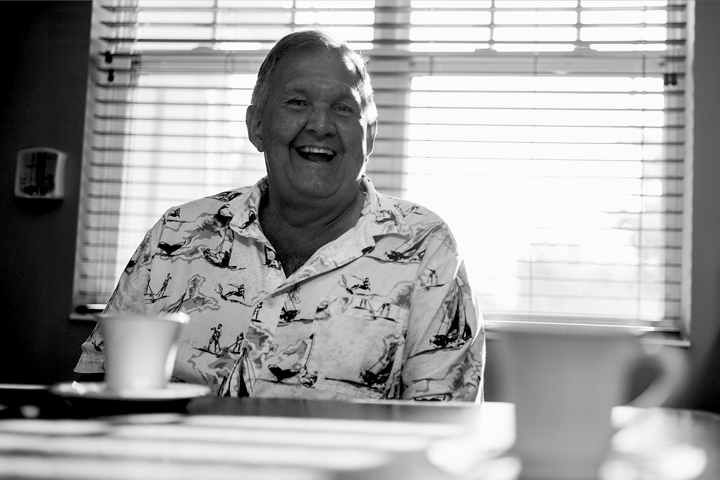 Black and white photo of a smiling older man wearing a button up beach shirt while sitting at a table with a cup of coffee