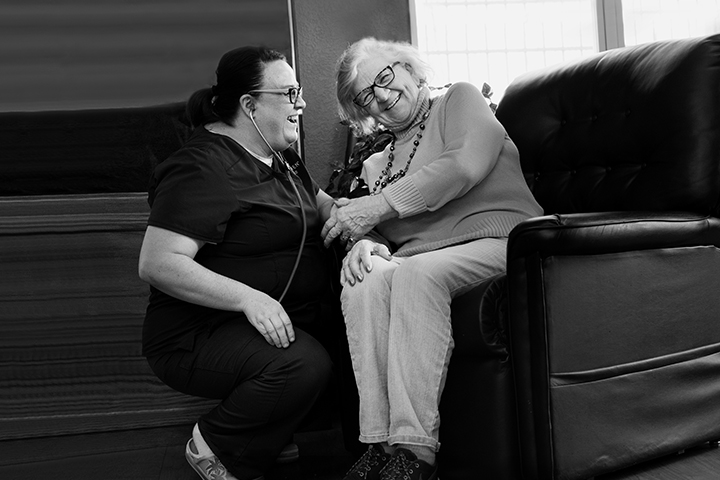 An older woman sits in a chair smiling as a caregiver kneels beside her, holding her arm, in a sunlit room. Black and white.
