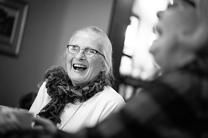 Black and white photo of an older women wearing a neck scarf and laughing with another women out of focus in the foreground