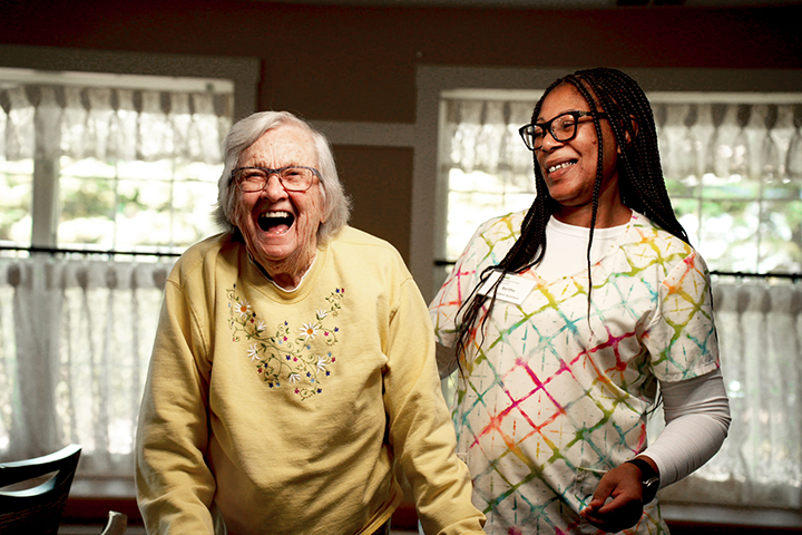 An older woman with a yellow sweatshirt laughs while a young, female team member assists her.