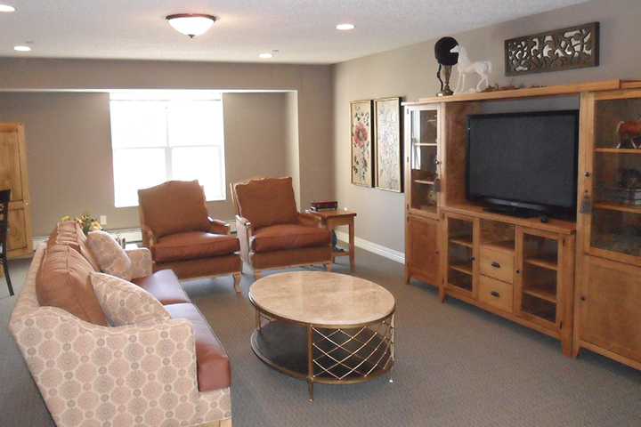 A carpeted lounge with sofa and upholstered chairs, coffee table, entertainment system, and a large window letting in lots of sunlight
