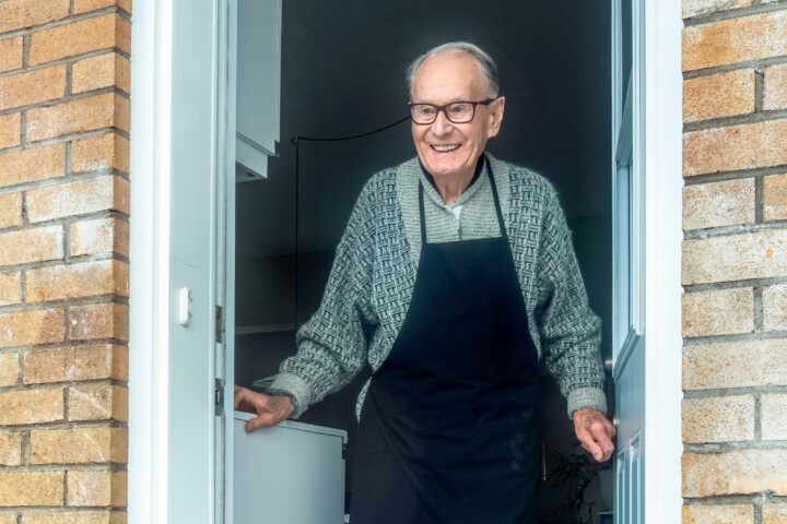 An older man wearing a cooking apron stands in the doorway of his home, smiling to greet a visitor.