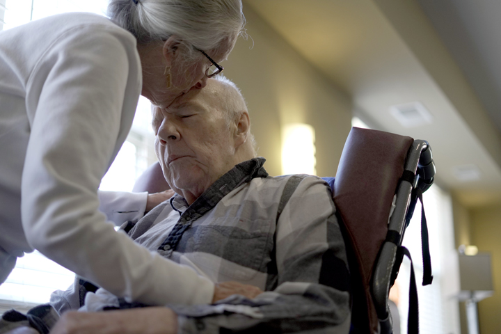 An older woman kisses the forehead of an older man in a wheelchair, showing tenderness and care.