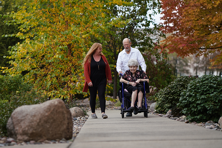 A young woman walking on a paved path next to an older gentleman who is pushing an older woman in a wheelchair.