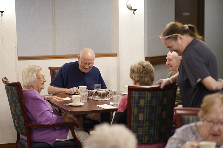 Four older people sit laughing around a dining table, while a dining server stands smiling beside them.