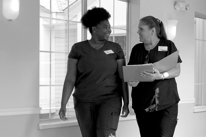 Black and white photo of two female caregivers walking in a hallway of large windows as they discuss the contents of a binder.