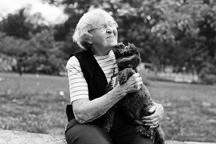 Black and white photo of an older woman kneeling down to embrace a small poodle as it licks her chin.