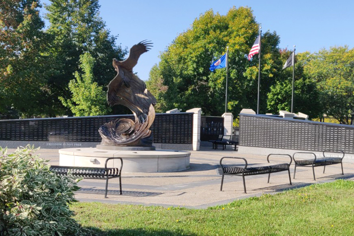 A large sculputure featuring a flying eagle is the centerpiece of a memorial with two converging walls of names beneath three flag poles: American flag, Minnesota flag, POW/MIA flag