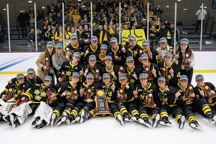 Post-game group photo of a women's hockey team with dark jerseys and gold trim, sitting and kneeling on the ice and holding the tournament trophy. Their fans cheer them on in the stands behind them.
