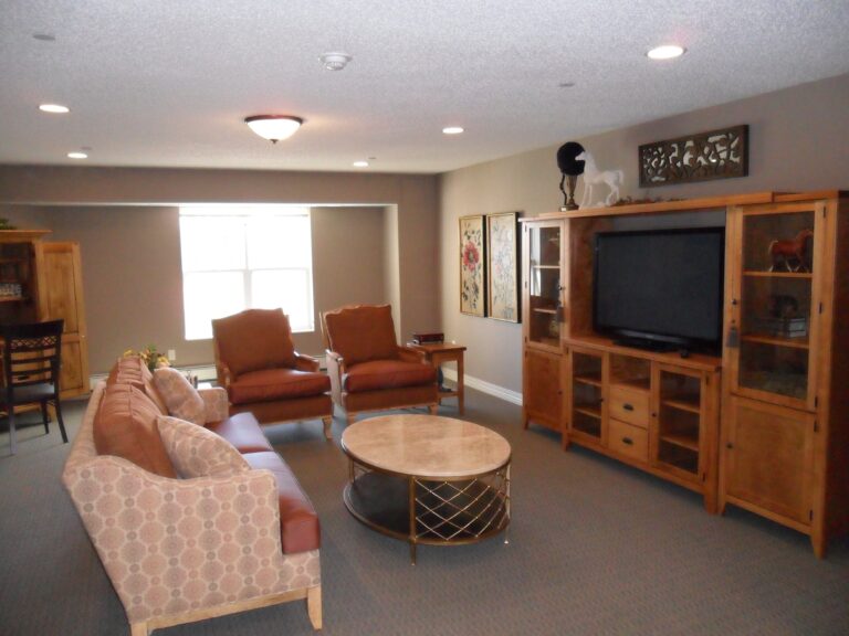 A carpeted lounge with sofa and upholstered chairs, coffee table, entertainment center, and a large window letting in lots of natural light.