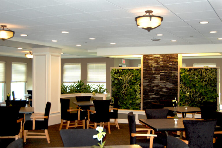 A bright dining room with four-top tables, upholstered arm chairs and a wall feature with living plants growing on the wall.