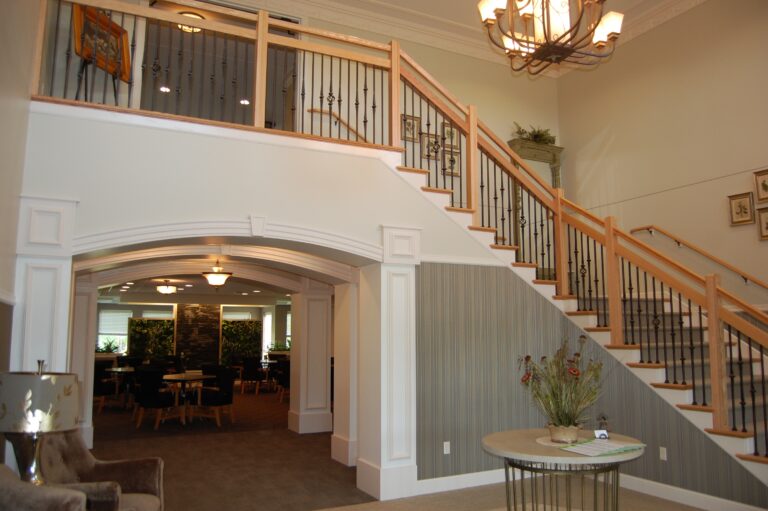 A chandelier hangs from the vaulted ceiling of a lobby with a large staircase and archway leading to a dining room.