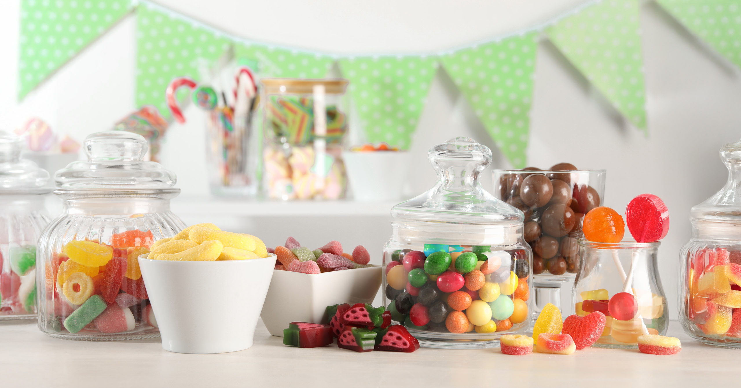 A candy buffet with glass jars filled with colorful sweets, a white bowl of yellow candies, and green bunting in the background