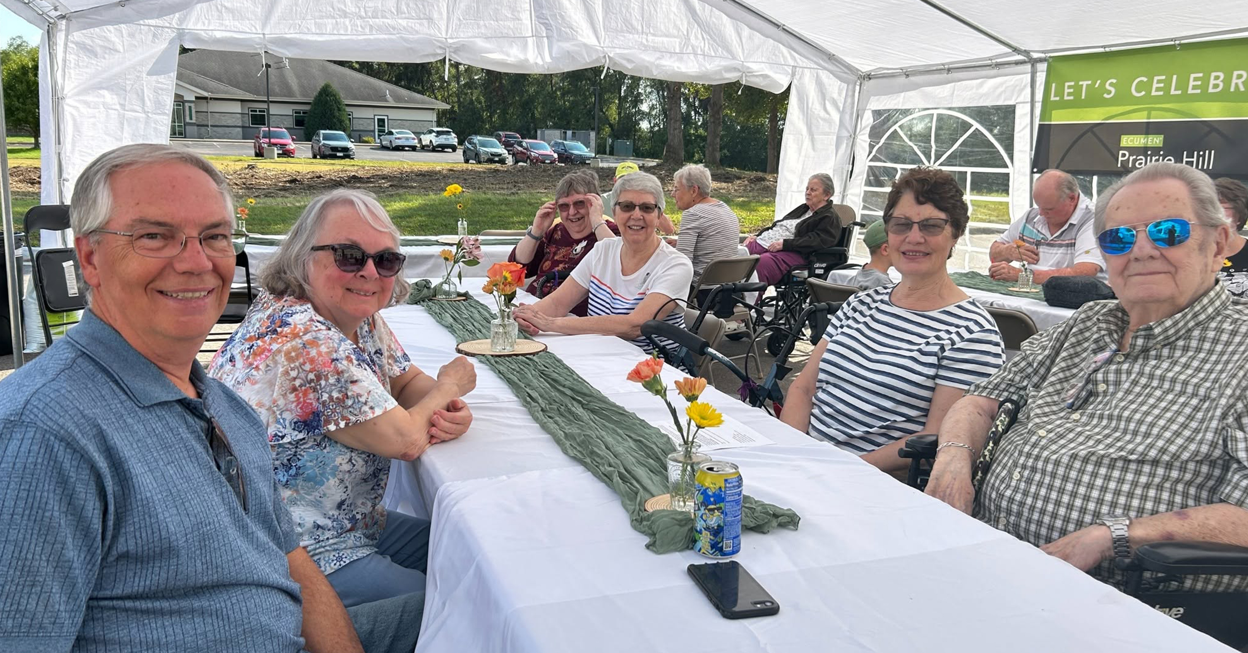 A group of seniors sits at long tables under a white tent during an outdoor picnic event.