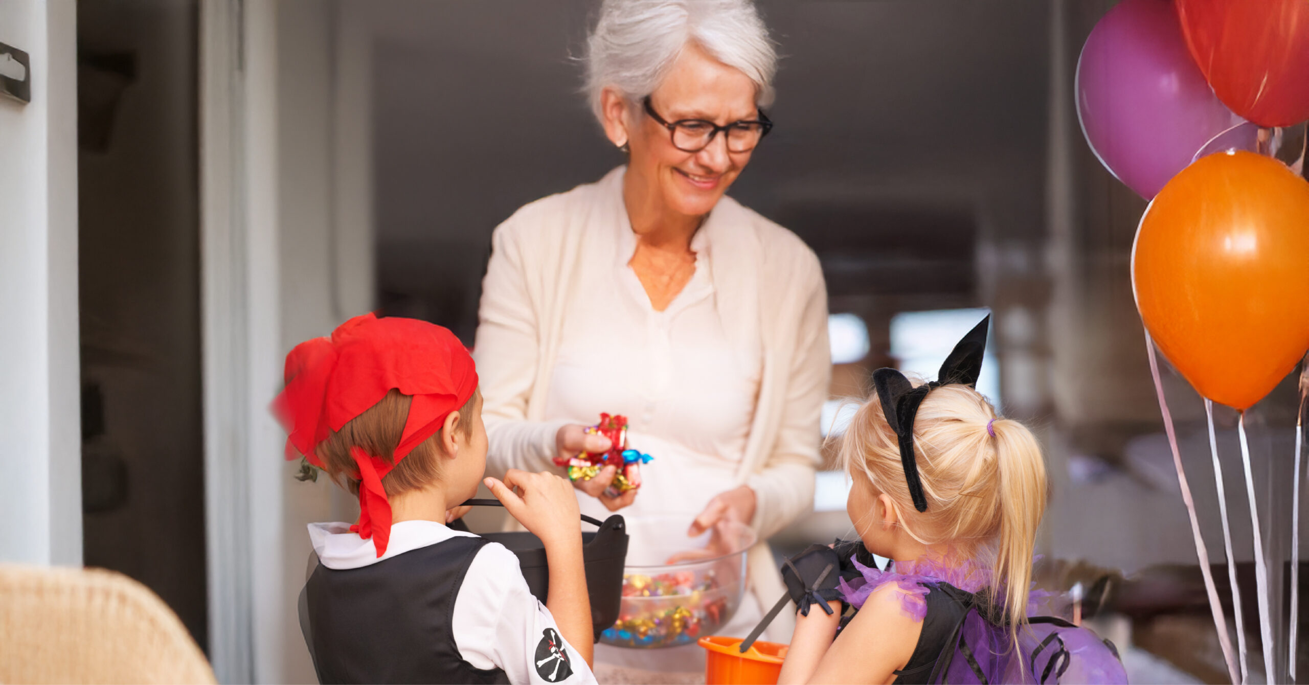An older woman with glasses hands candy to two children in Halloween costumes, with balloons.
