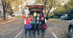 Three children in Halloween costumes stand in front of a car decorated for a trunk-or-treat event, holding candy buckets on a leaf-covered street.